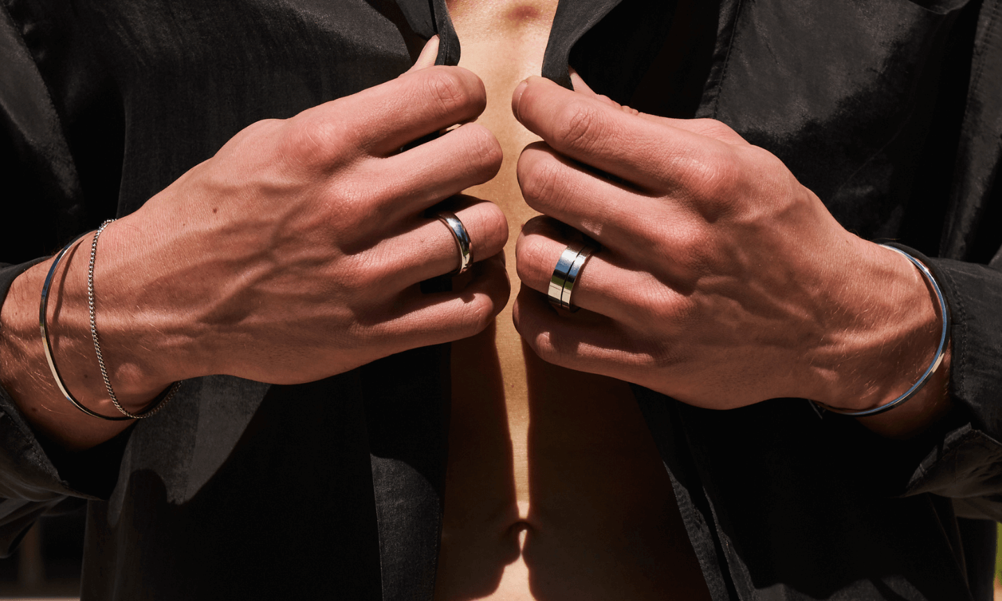 Man wearing a classic open collar black shirt and styling essential silver jewelry including rings, bracelets and bangles