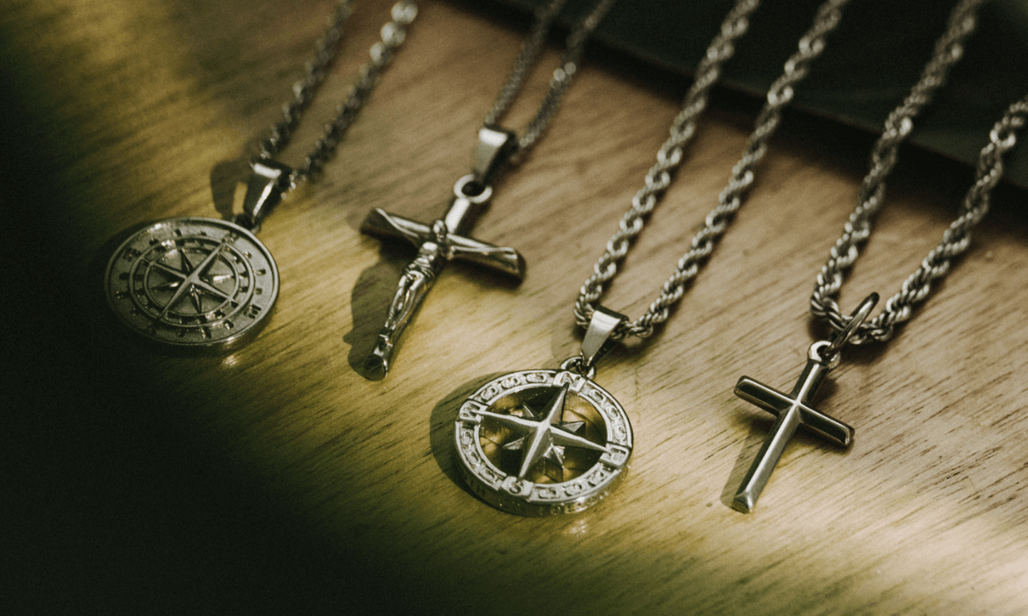 Four men’s silver pendants laid on a wooden surface, including two cross designs and two compass designs, each suspended from sterling silver chains. The lighting highlights the polished finish and rugged detail of each piece.