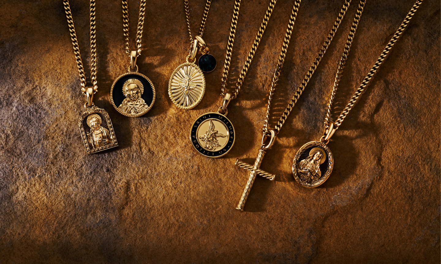 A row of men's 18K gold and gold vermeil pendant necklaces, including religious medallions, a Saint Michael coin and a cross, laid across a warm stone surface.