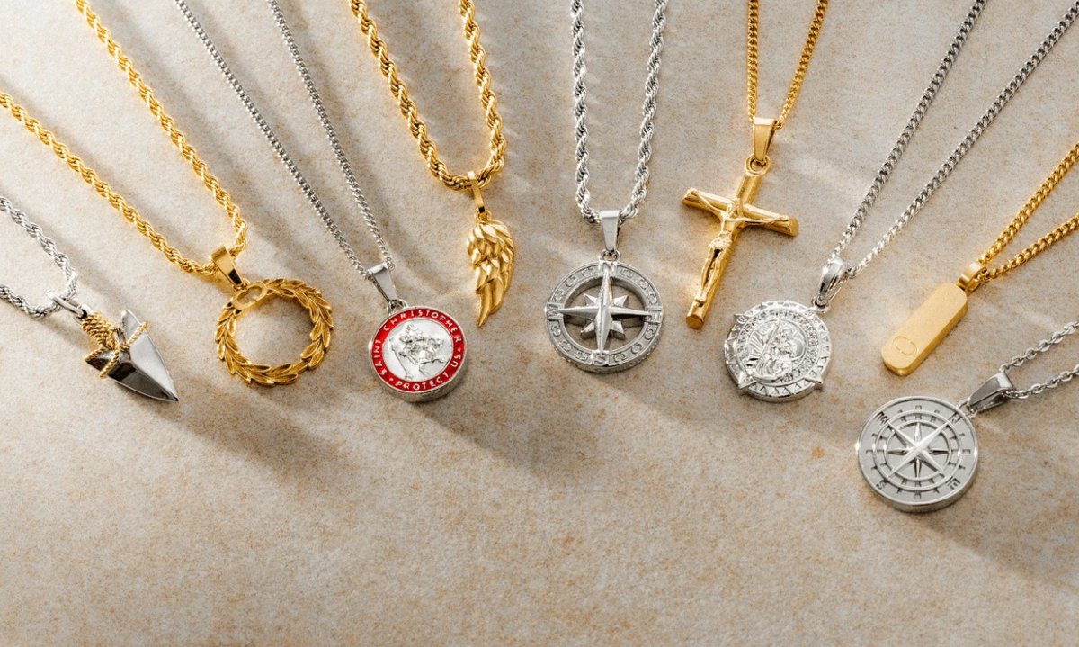 Flat lay of mixed gold and silver pendant necklaces, including compass designs, crosses, angel wings, and laurel wreaths. Each piece reflects a different meaning like faith, direction, strength.