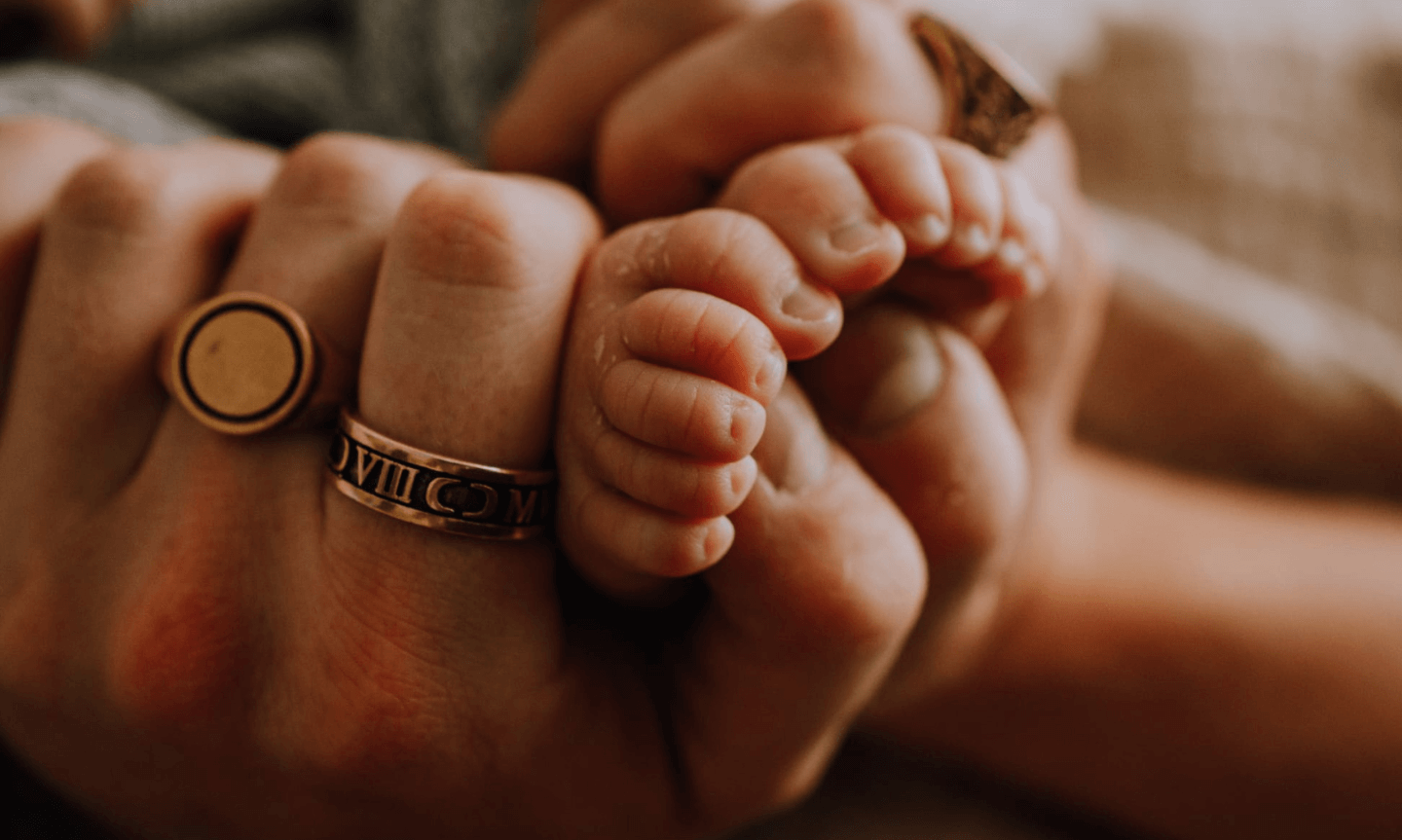 Man's hands wrapped around a babies foot. He's wearing a gold signet ring and band ring. The Number One Men's Jewelry Gift for Fathers-to-Be.